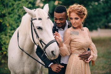 Newlyweds are standing near a white horse in nature