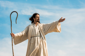 Jesus with outstretched hand holding wooden cane against blue sky