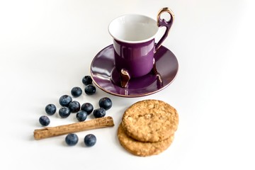 Purple vintage tea cup , blueberries, cinnamon stick and cookies against white background. Breakfast, food and infusion concept.