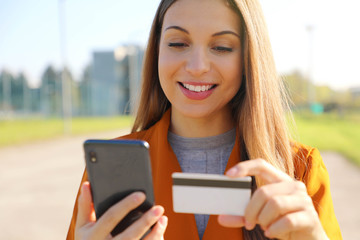 Close up of beautiful woman reading credit card number doing online payment on her smart phone outside on a sunny autumn day.