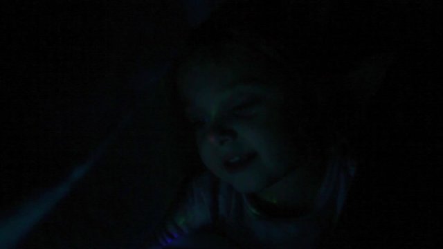 Little female toddler plays under the covers with a multicolored lamp.  The little girl sings and talks while having fun.