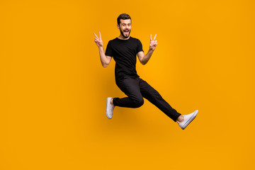 Fototapeta na wymiar Full length photo of handsome excited guy jumping high raising hands showing v-sign symbols greeting friends meet street wear black t-shirt pants isolated yellow color background