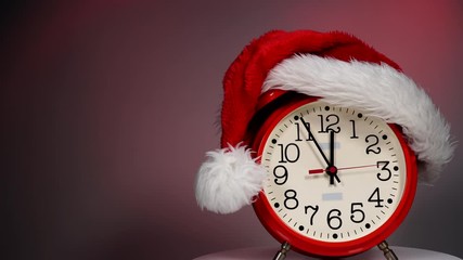 Christmas time concept. Santa Claus hat and big red clock ticking showing twelve hours, on dark red background. Waiting for celebration. Time lapse