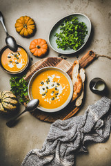 Autumn, winter warming seasonal meal. Flat-lay of pumpkin soup with seeds, parsley, cream and bread in bowls over grey concrete background, top view. Vegan, vegetarian, healthy, comfort food concept