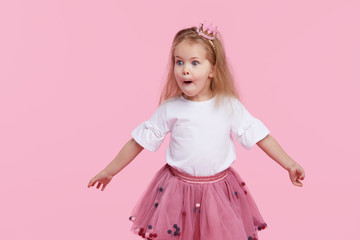 A cheerful little girl with in a tulle skirt and princess crown on her head isolated on a pink background. Celebrating a vibrant carnival for kids, birthday party, having fun 