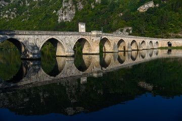 Fototapeta premium The Ottoman Mehmed Pasa Sokolovic Bridge in Visegrad, Bosnian mountains, with fantastic sky scape and river reflection. Bosnia and Herzegovina.