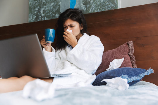 Photo of ill woman sneezing and drinking tea while using laptop in bed