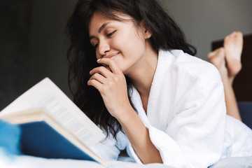 Photo of satisfied woman wearing housecoat reading book in bed at home