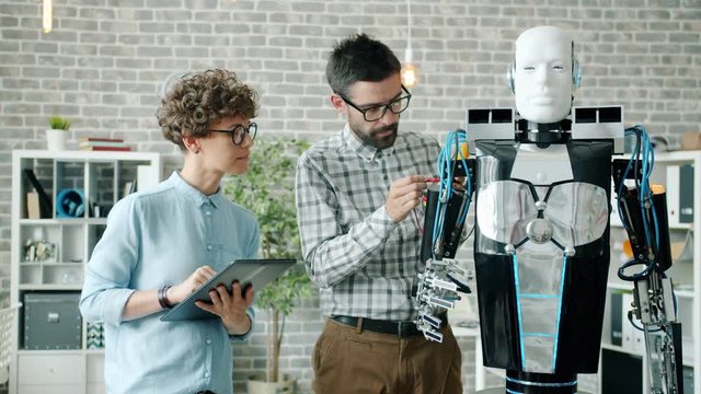 Man and woman testing modern human-like robot in laboratory using tablet indoors. Computers, artificial intelligence and robotics engineering concept.