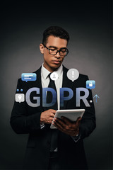 african american businessman in glasses using digital tablet on dark background with gdpr illustration