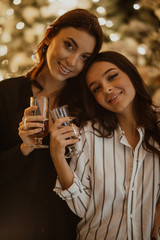 Lesbian couple holds glasses of wine against background of Christmas decorations.