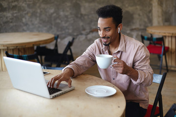 Fototapeta na wymiar Cheerful young bearded man with dark skin working in coffee house with modern laptop using public wi-fi, reading mail and having good day, keeping cup of tea in his hand and smiling