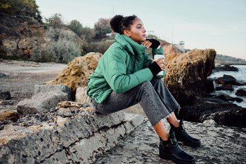 Side view of beautiful casual African American girl in down jacket with cup to go dreamily sitting on stones by the sea