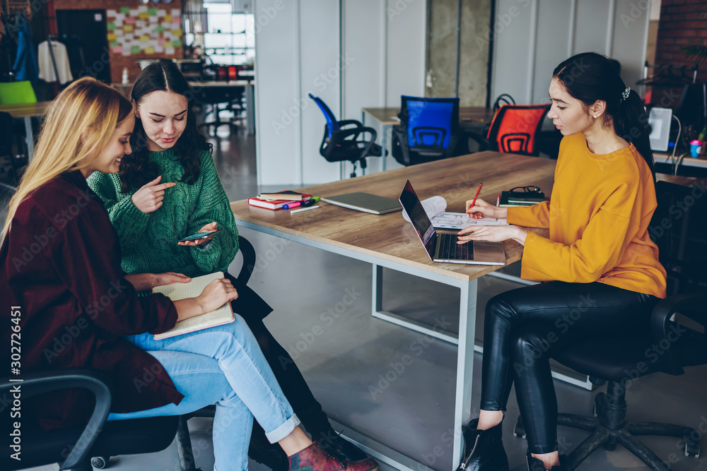 Aufkleber smiling hipster girls having conversation on break while other colleague using laptop computer for w
