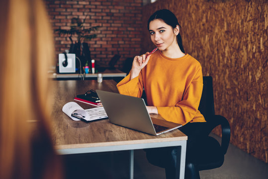Portrait of beautiful brunette young woman dressed in casual yellow sweater looking at camera while working on new graphic at modern laptop computer connected to wireless 4G iternet sitting in office