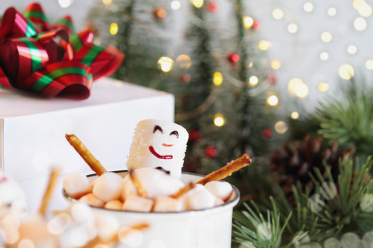 Two snowmen soaking in a hot cup of cocoa surrounded by mini marshmallows. Extreme selective focus on snowman's face with blurred foreground and background. Christmas trees and gift in background.