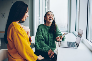 Overjoyed female laughing at funny content from network sitting next to colleague using modern...