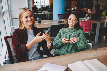 Joyful caucasian hipster girls having fun together in coworking space using mobile phone and bluetooth connection for share multimedia, happy cheerful female friends laughing at photos on cellulars