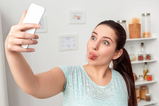 Body Care. Chubby girl standing in kitchen taking selfie on smartphone showing tongue to camera playful close-up