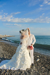 Fototapeta na wymiar Wedding by the sea. Beautiful couple of newlyweds. Bride with a red bouquet in a wedding dress at the water. Bride and groom kissing on stone beach during sunrise time. Just married in Batumi, Georgia