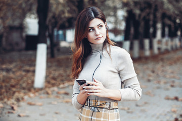 Image of beautiful stylish woman holding mobile phone. Young european girl standing at the street and using cellphone. Woman listening to music with the phone and having fun. Mobile internet concept. 