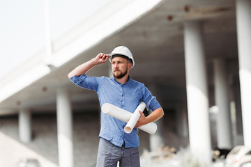 The foreman in a white protective helmet holds the drawings in his hands, against the background of the span of the bridge under construction and looks away