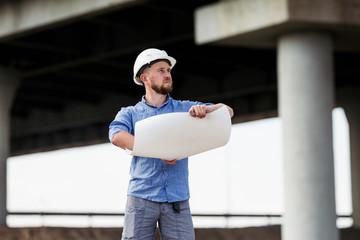 A foreman in a white protective helmet examines the drawings on the background of the span of the bridge under construction
