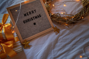 Felt letter board Merry Christmas on the bed decorated with golden wreath, lights and gift boxes