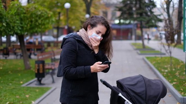 Young mother with a stroller in the park in a medical mask speaks on the phone in the fall.