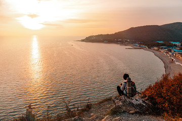 Fototapeta premium Tourism and sports recreation. A young woman with a tourist backpack on her back sits on a rock and looks at the sunset and the sea. View of the Bay. Copy space