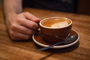 Male fingers holding brown cup of cappuccino on wooden table, Blurred background.