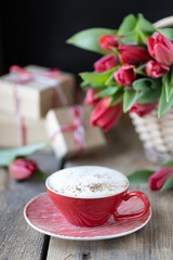 Red cup of coffee close-up, a bouquet of red tulip and gift boxes on wooden background