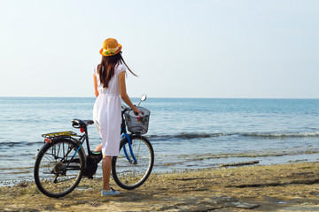 Girl in a white dress with a bicycle on the seashore, back view.