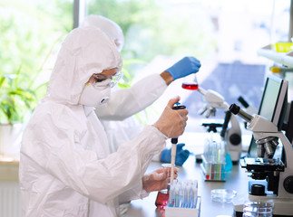 Scientists in protection suits and masks working in research lab using laboratory equipment: microscopes, test tubes.