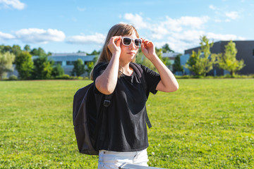 Happy/smiling teen girl/student standing in front of a high school and sports field while wearing sunglasses.