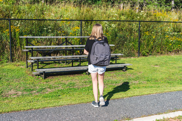 Teen girl walking to a set of bleachers near her school while wearing a backpack and holding...