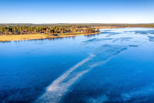 Aerial scenic view over Swedish village and crystal clear frozen lake in Northern Scandianvia. Blue ice, clear skies, sunny winter day. Frozen lake like mirror, Umea city, Vasterbotten, Sweden