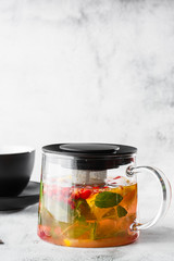 Glass teapot of cranberry, orange and mint or yellow tea on black cup isolated on bright marble background. Overhead view, copy space. Advertising for cafe menu. Coffee shop menu. Vertical photo.