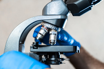 Male laboratory assistant examining biomaterial samples in a microscope. Cllose up hands in blue rubber gloves adjust microscope