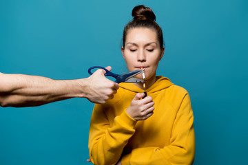 a girl in bright clothes is going to light a cigarette, but a male hand with scissors is going to stop her