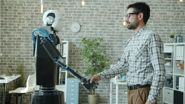 Young male developer and manlike robot cyborg are shaking hands standing in office together, person is enjoying artificial intelligence. People and innovation concept.