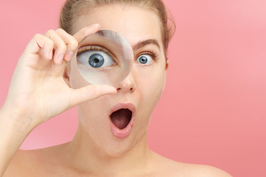 Fototapeta Young woman looking at the camera through magnifier with funny surprised expression, closeup isolated on pink. Enlarged eye, optical illusion, search