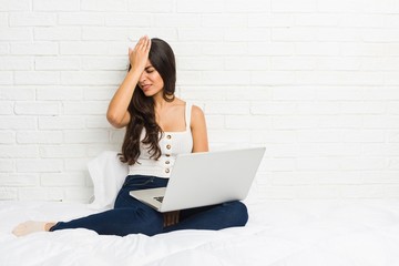 Young arab woman working with her laptop on the bed forgetting something, slapping forehead with palm and closing eyes.