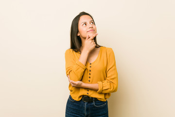 Young hispanic woman against a beige background looking sideways with doubtful and skeptical expression.