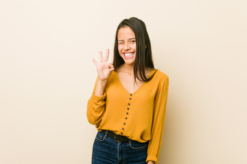 Young hispanic woman against a beige background winks an eye and holds an okay gesture with hand.
