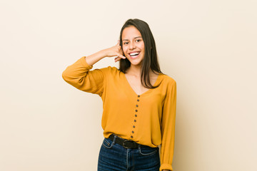 Young hispanic woman against a beige background showing a mobile phone call gesture with fingers.