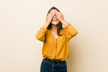 Young hispanic woman against a beige background covers eyes with hands, smiles broadly waiting for a surprise.