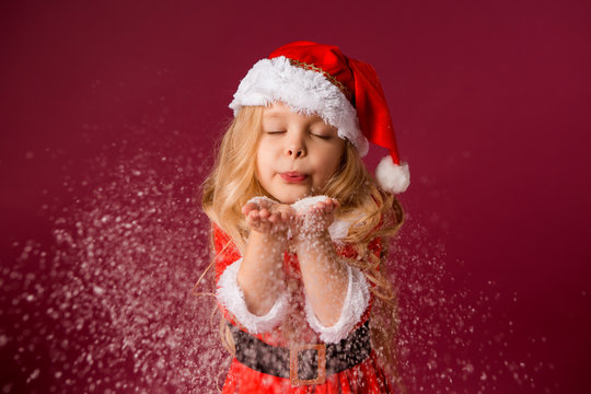 little blonde girl in a Santa suit blows snow off her hands. red background isolate. space for text