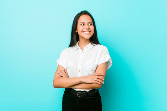 Young hispanic cool woman against a blue wall smiling confident with crossed arms.