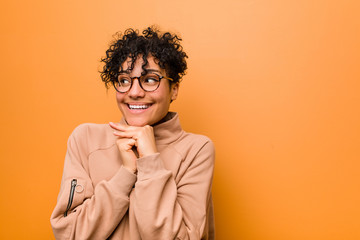 Young mixed african american woman against a brown background keeps hands under chin, is looking happily aside.